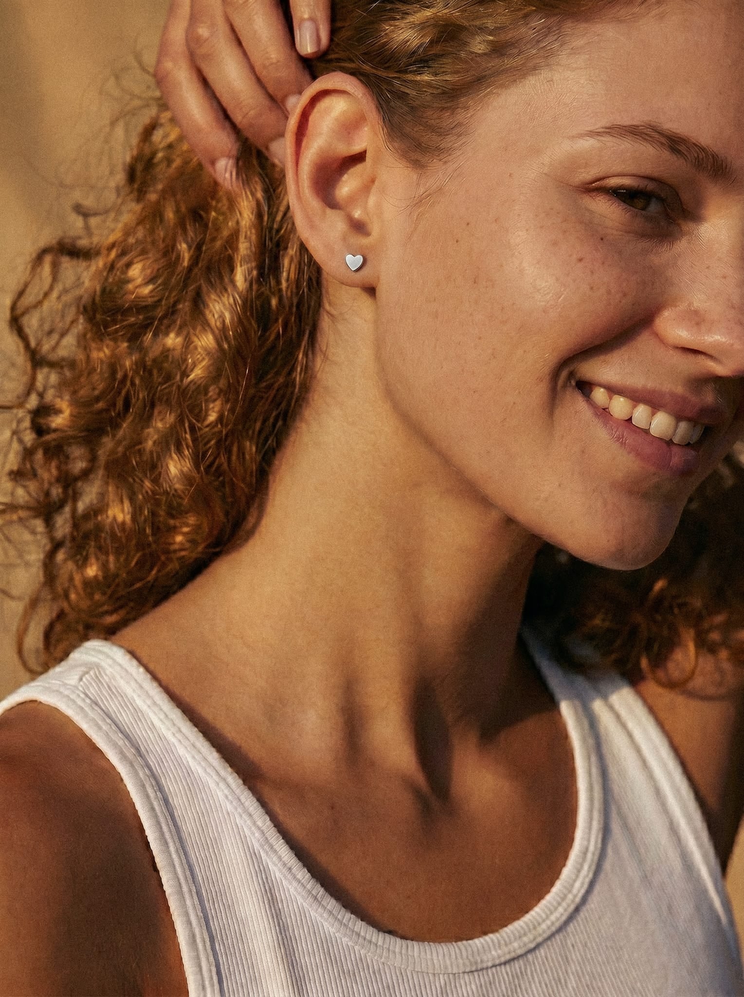 Una mujer de pelo rizado sonríe bajo la cálida luz del sol; lleva una camiseta sin mangas blanca y unos pendientes de botón en forma de corazón «LOVE LISO SILVER» de Titanio-Noble Art, con la mano levantada hacia la cabeza, lo que deja al descubierto un delicado piercing.