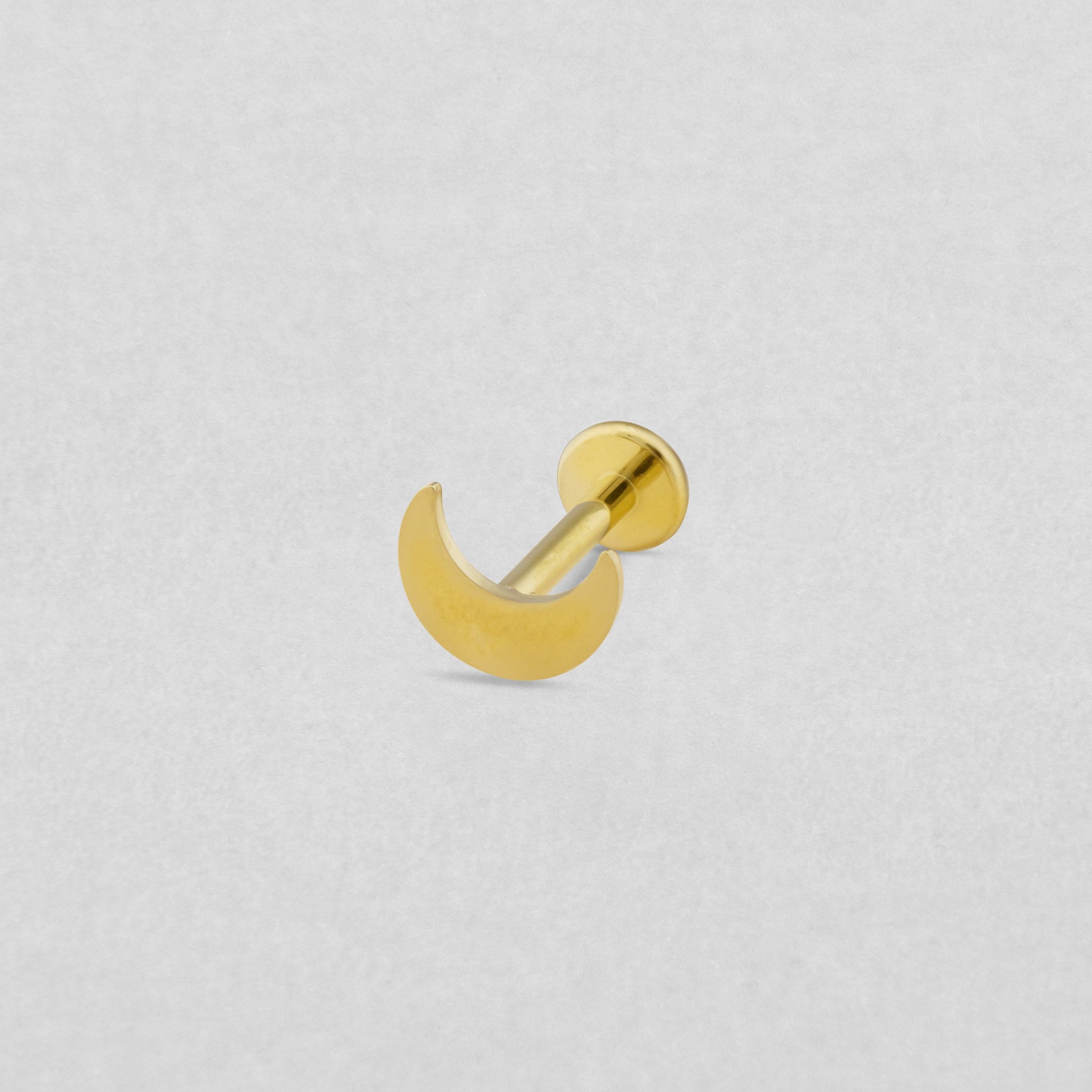 El modelo «Luna liso» de Titanio-Noble Art es un pendiente de botón con forma de media luna dorada y parte trasera plana, ideal para los amantes de los piercings, que se presenta sobre un fondo gris claro con textura.
