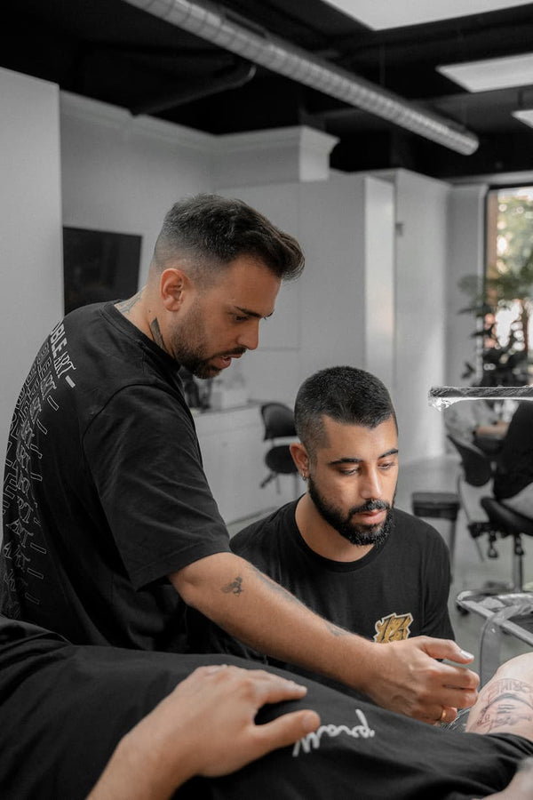 Dos hombres en un moderno estudio especializado en tatuajes: uno de ellos está utilizando una máquina de tatuar en el brazo de un cliente, mientras que el otro le ayuda. El ambiente profesional sugiere que cuentan con experiencia tanto en tatuajes como, posiblemente, en servicios de piercing o de eliminación con láser.